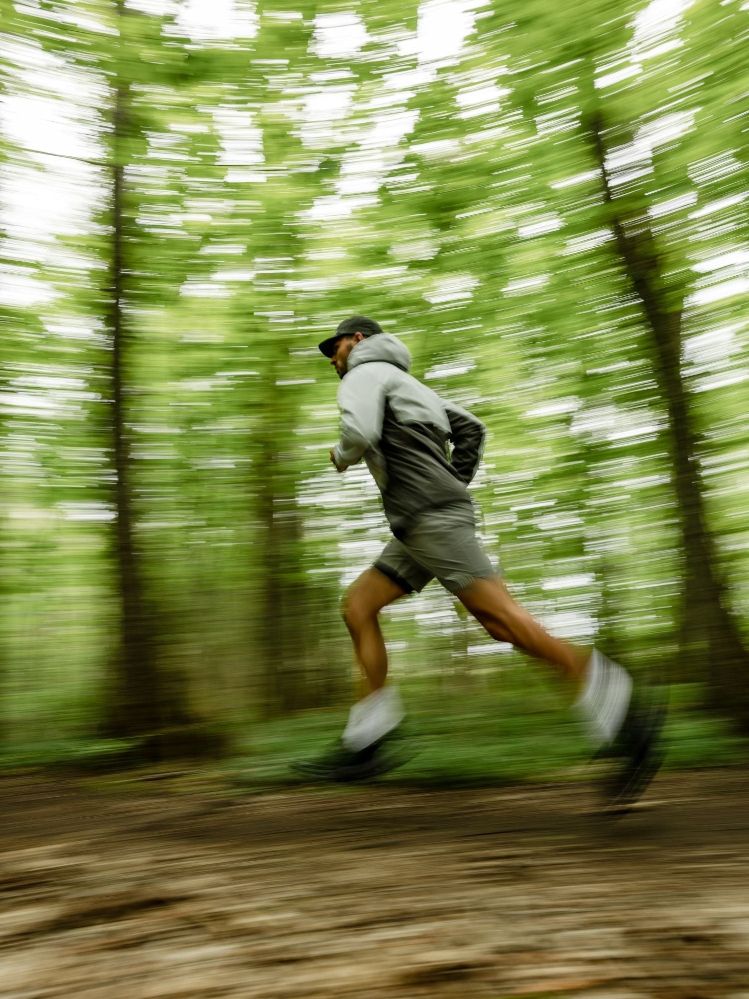 Eine Person in sportlicher Kleidung läuft schnell durch einen grünen Wald. Der Hintergrund ist unscharf, was die Bewegung und Geschwindigkeit betont. Der Läufer trägt einen Kapuzenpulli, kurze Hosen, eine Mütze und einen kleinen Rucksack.