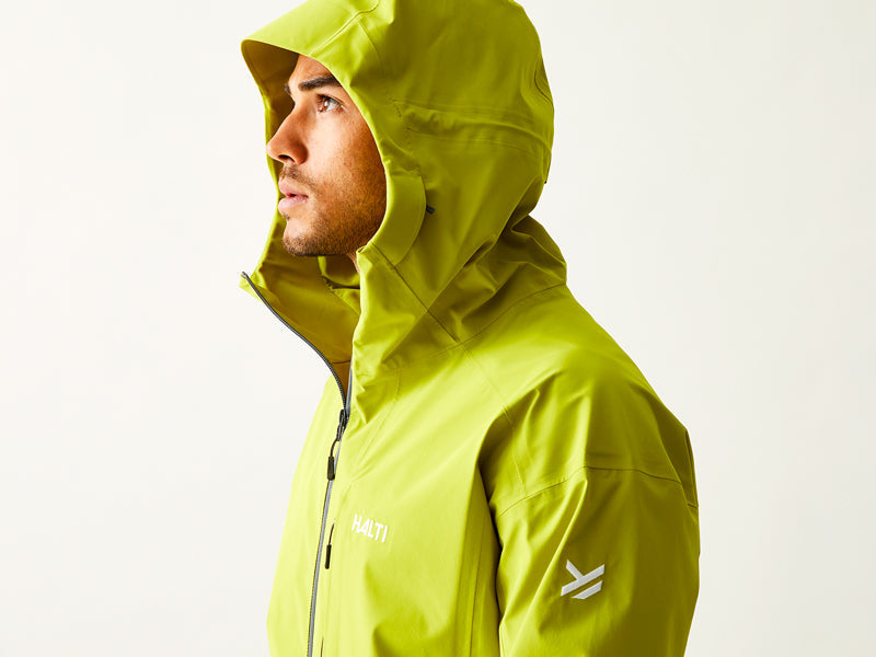 Eine Person in einer orangefarbenen Halti-Jacke steht im Freien vor einem felsigen Hintergrund; in einem Bild sind zwei Personen zu sehen, die am Wasser wandern. Text: Getestet für das Unerwartete, EST. 1976, HALTI.COM, und HALTI.