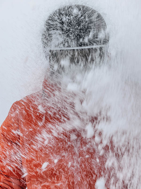 Zwei Personen in Winterkleidung stehen bei Sonnenuntergang mit Wanderstöcken und Rucksäcken auf einem verschneiten Berg. Im Vordergrund weht der Schnee, und im Hintergrund sind ferne Berge zu sehen.