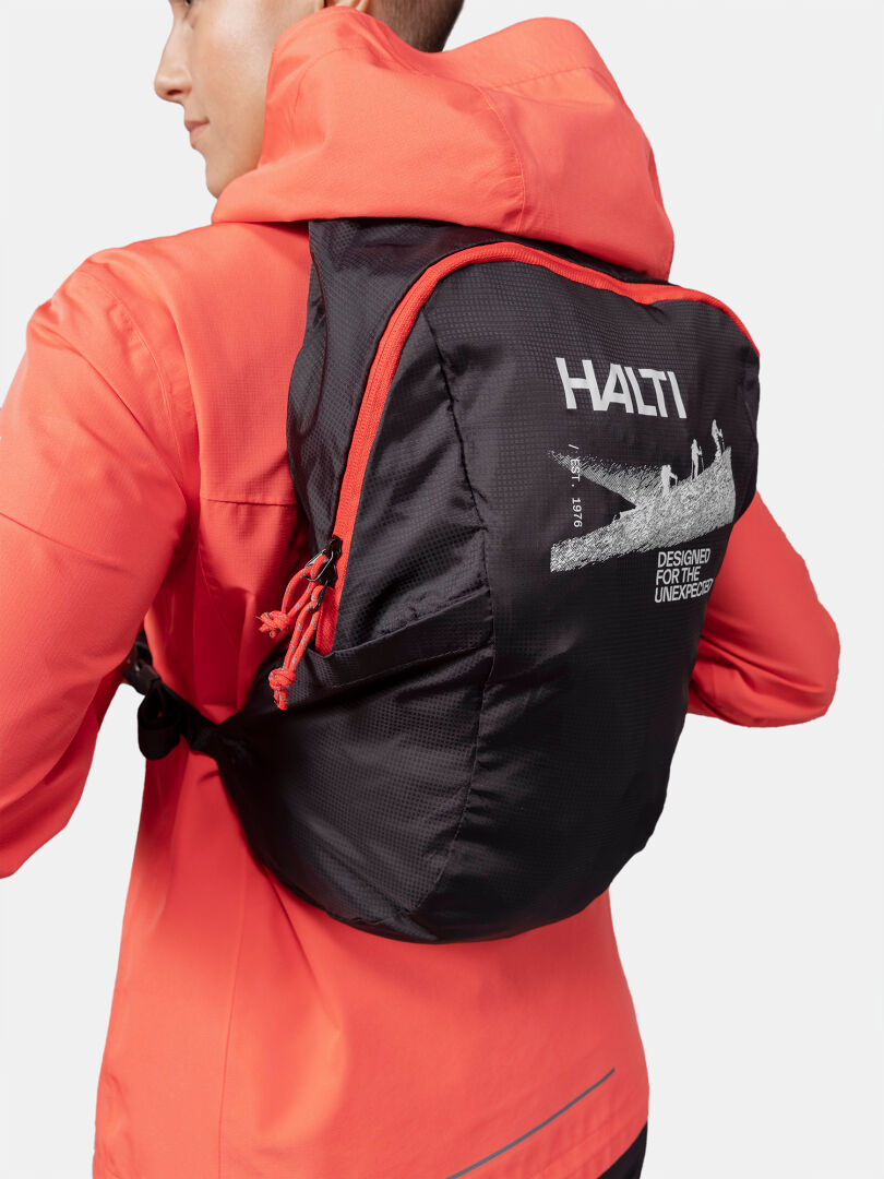 Person in leuchtend orangefarbener Jacke trägt einen schwarzen Halti Streetpack Rucksack mit weißer Grafik auf dem Rücken.