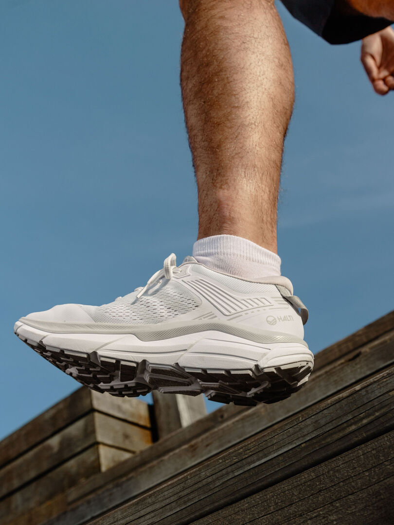 Eine Person in Halti Tempo 3 Sneaker Herren und weißen Socken steht unter einem klaren blauen Himmel auf einer Holzfläche.