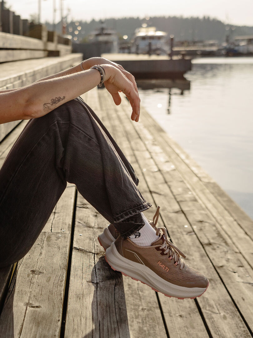 Eine Person in dunklen Hosen und beigen Halti Rapid Sneaker Damen sitzt auf einem Holzsteg am Wasser, dahinter sind Boote.