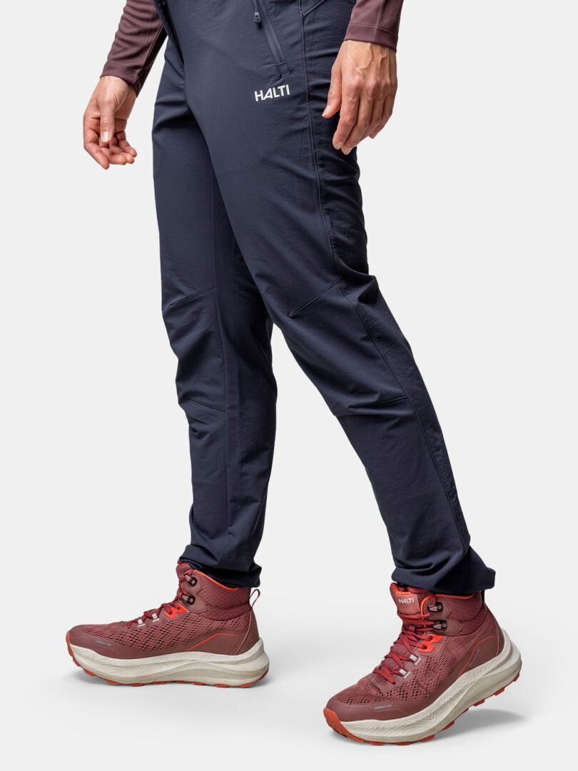 Frau in dunkelblauer Halti Hose und Halti Rapid DX Mid Outdoor Schuhe Damen steht vor einem schlichten hellen Hintergrund.