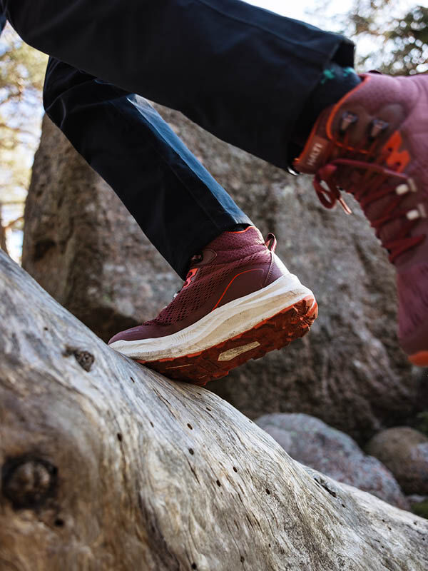 Eine Frau in Halti Rapid DX Mid Outdoor Schuhen klettert auf einen schrägen Baumstamm; im Hintergrund sind Felsen und Natur zu sehen.