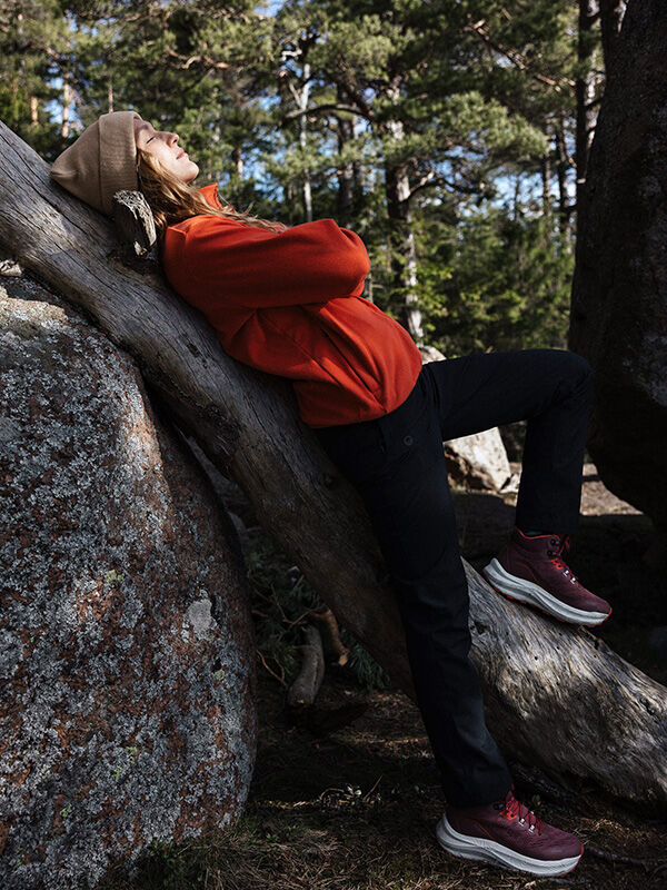 Eine Person in einer roten Jacke, einem beigen Hut und Halti Rapid DX Mid Outdoor-Schuhen für Damen lehnt an einem umgestürzten Baum im Wald.