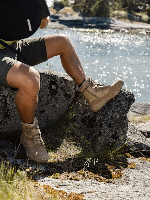 Eine Person in Halti Regis DX Stiefel Herren und Shorts sitzt an einem sonnigen Tag auf einem Felsen am Wasser.