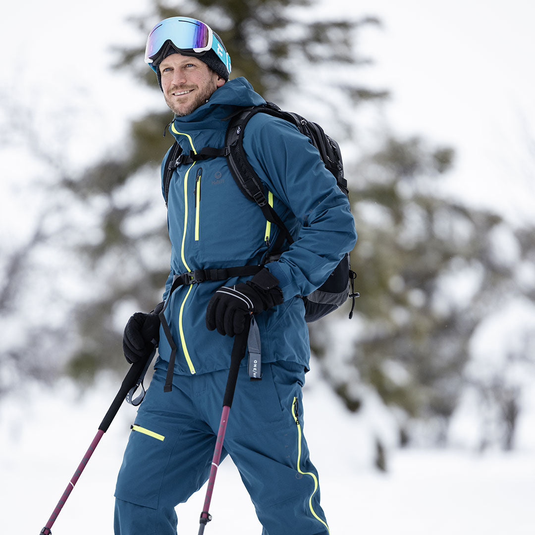 Eine Person in blauer Skibekleidung und mit Skibrille trägt Halti Alpine 3L DX Skihose Herren und steht mit Skistöcken im Schnee.