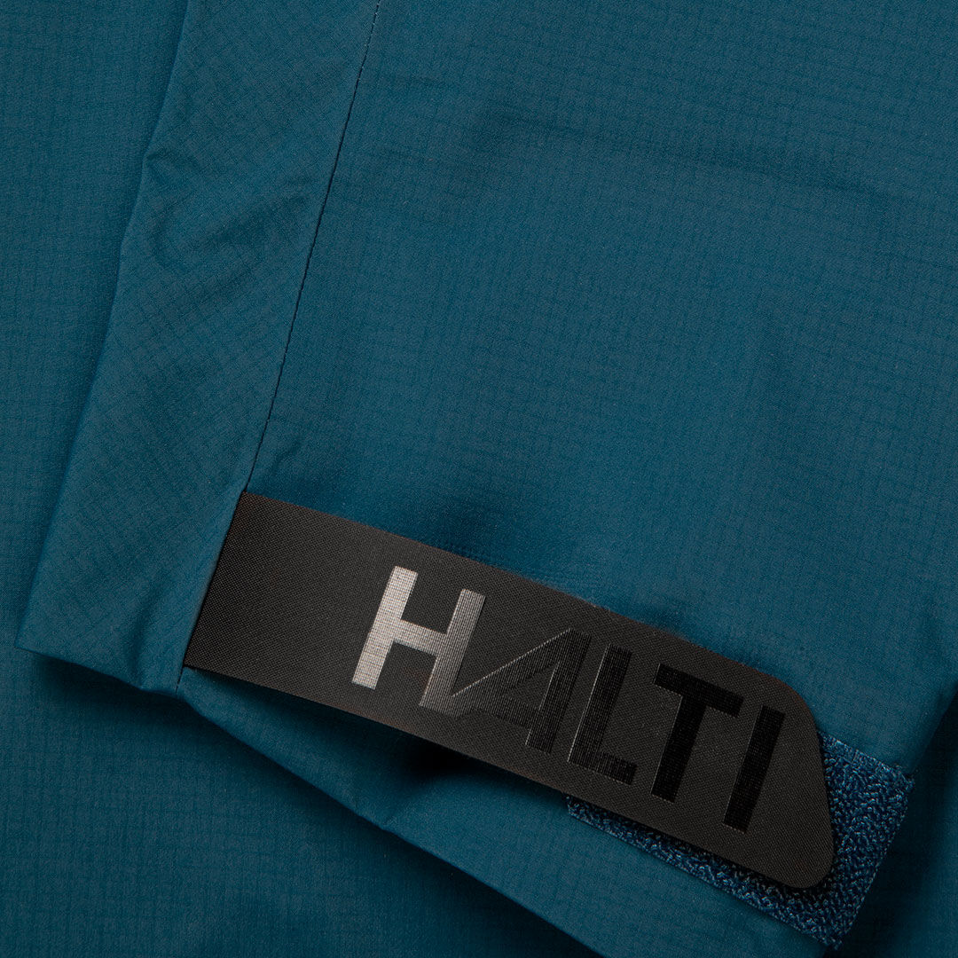 Nahaufnahme des blauen Stoffs und des schwarzen Riemens mit Halti-Logo, Teil der Alpine 3L DX Skijacke Unisex.