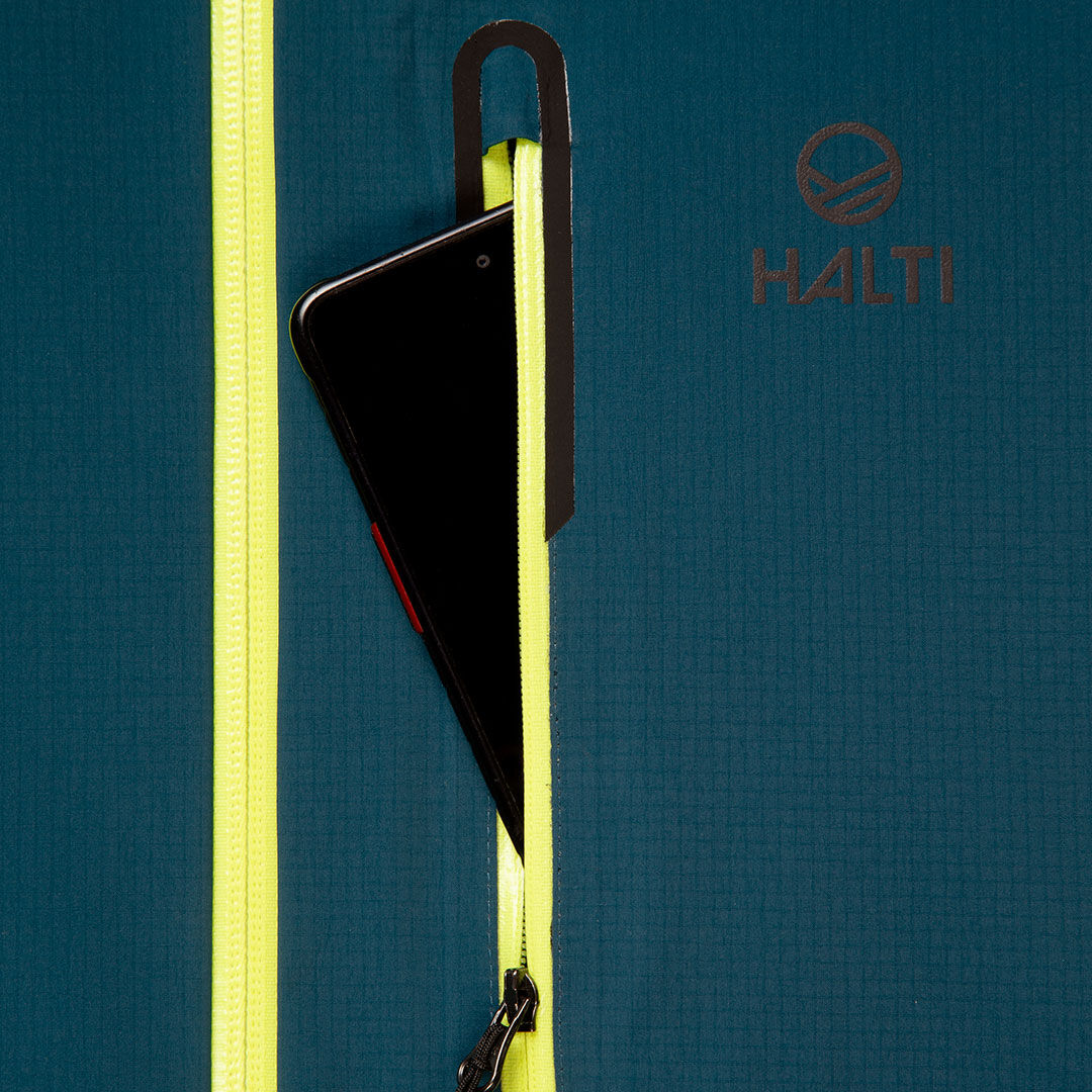 Ein schwarzes Smartphone in der Reißverschlusstasche einer tealfarbenen Halti Alpine 3L DX Skijacke Unisex mit gelben Reißverschlüssen.