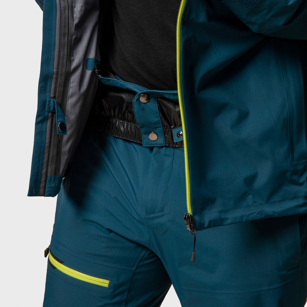Person in einer blauen Halti Alpine 3L DX Skijacke Unisex und passender Hose mit gelben Reißverschlüssen; die Jacke ist am Bund offen.