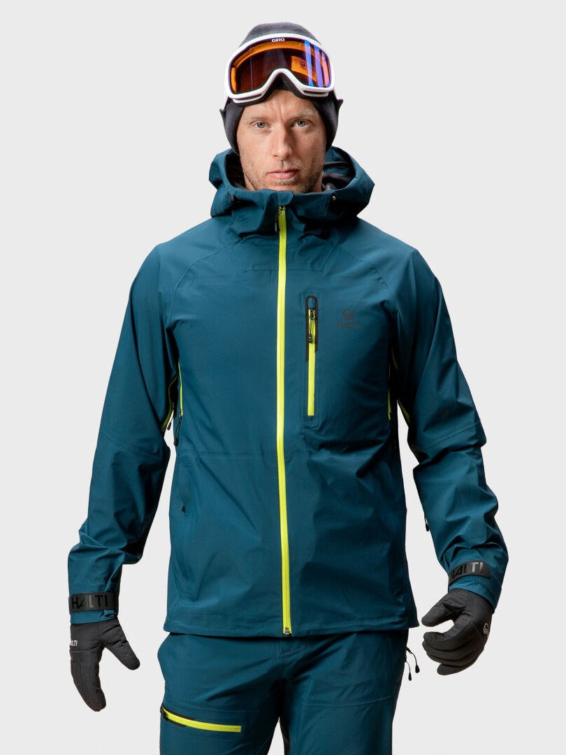 Eine Person in blauer Skikleidung trägt die Halti Alpine 3L DX Skijacke Unisex, bereit zum Skifahren im Hinterland.
