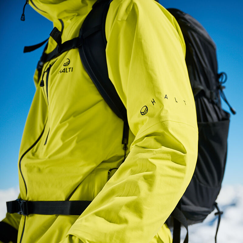 Person trägt Halti Alpine 3L DX Skijacke Unisex in leuchtendem Gelb mit einem schwarzen Rucksack vor einem blauen Himmel.