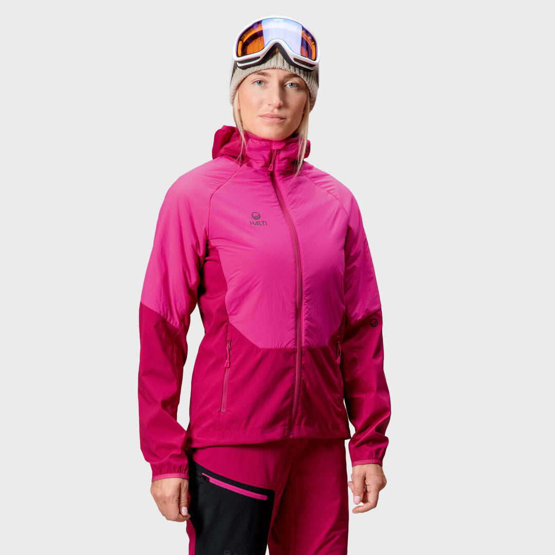 Person in einer leuchtend rosa Halti Crust Damen Layer Jacke, passender Hose, Skibrille und Mütze auf schlichtem Hintergrund.