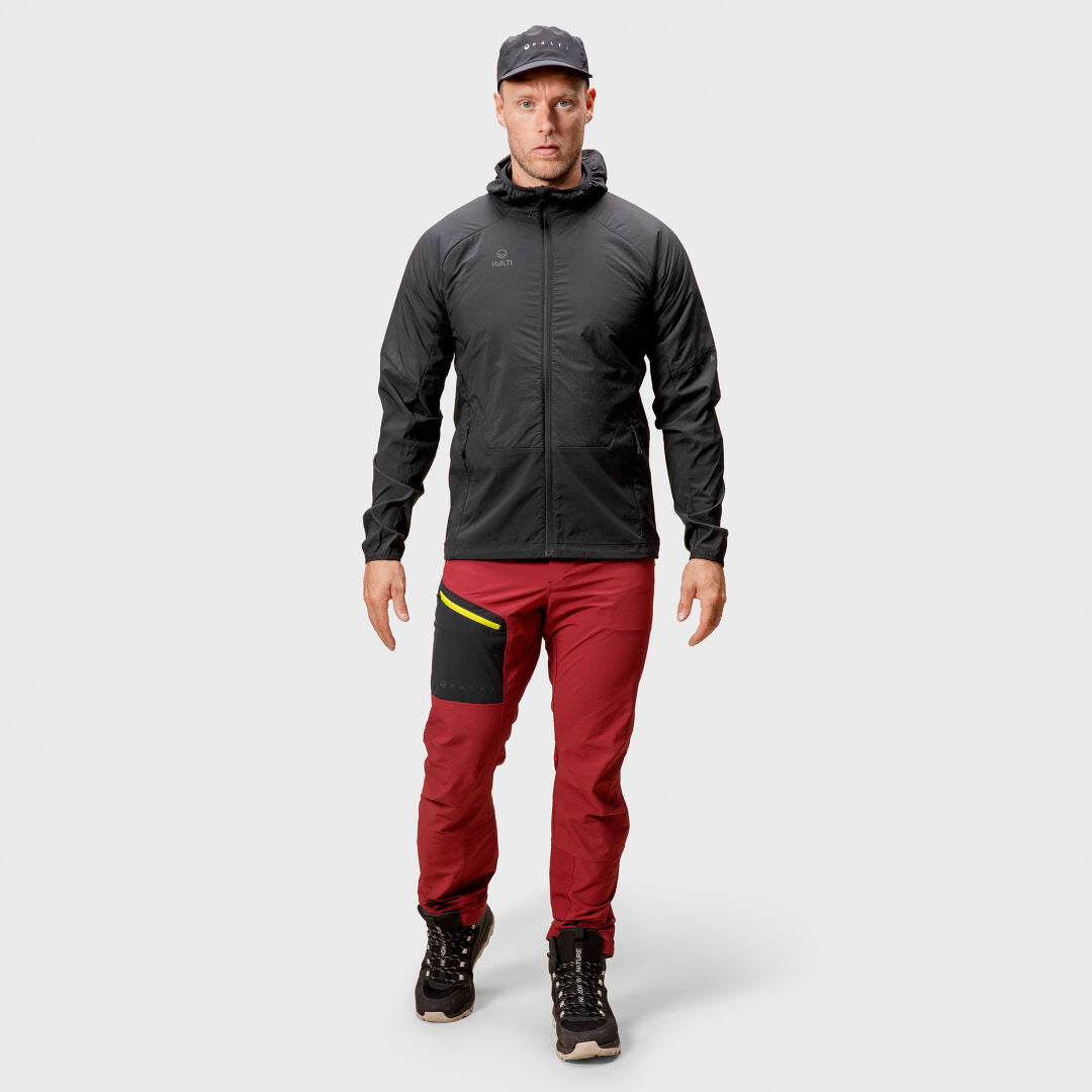 Eine Person trägt eine Halti Crust Herren Layer Jacke, rote Hosen mit schwarzem Flicken, schwarze Schuhe und eine Mütze und blickt nach vorne.