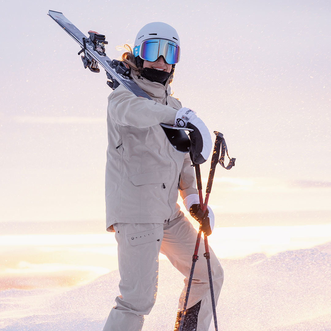 Frau in beiger Halti Settler 3L DX Ski Jacke Damen steht mit Skiern im Schnee, gewärmt durch DrymaxX Nano tech unter einem strahlenden Himmel.