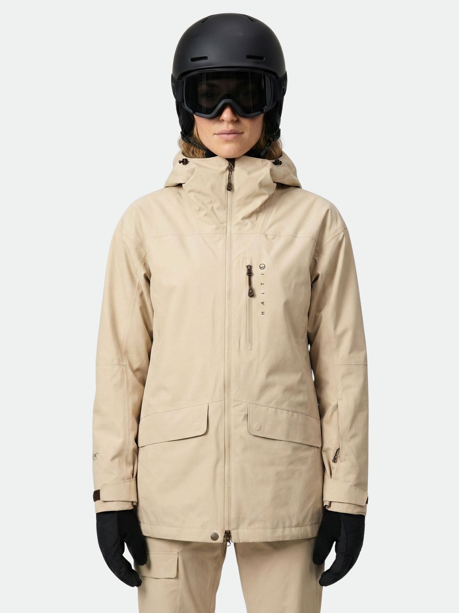 Eine Person in einer beigen Halti Settler 3L DX Ski Jacke Damen und Helm blickt nach vorne vor einem schlichten hellen Hintergrund.