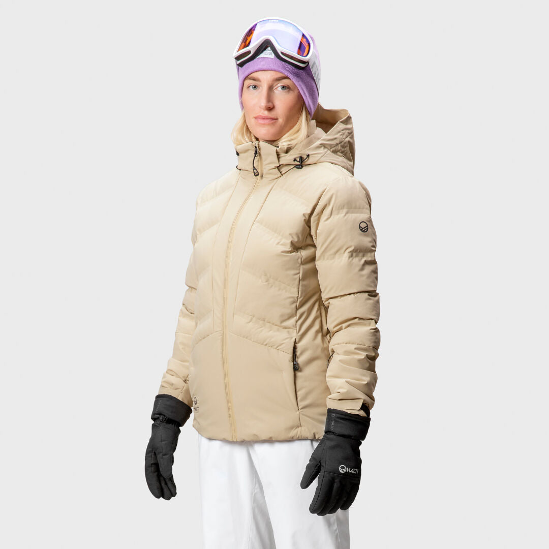 Eine Person in einer Halti Nordic Arcty Skijacke für Damen, weißen Hosen, schwarzen Handschuhen, einer lila Mütze und einer Skibrille.