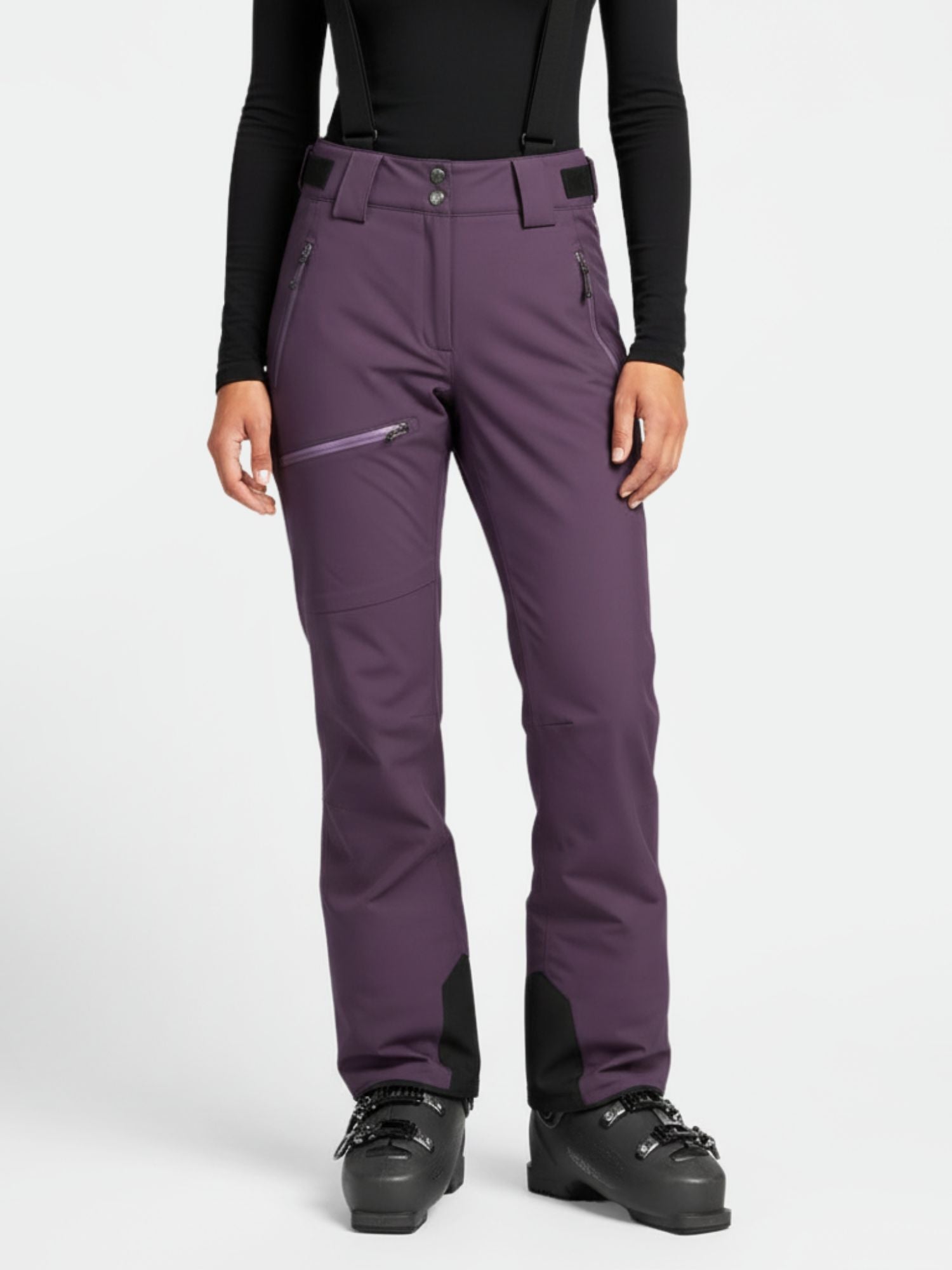 Eine Person in einer lila Halti Carvey DX Skihose Damen mit schwarzen Hosenträgern, schwarzen Stiefeln und einem schwarzen Oberteil steht aufrecht.