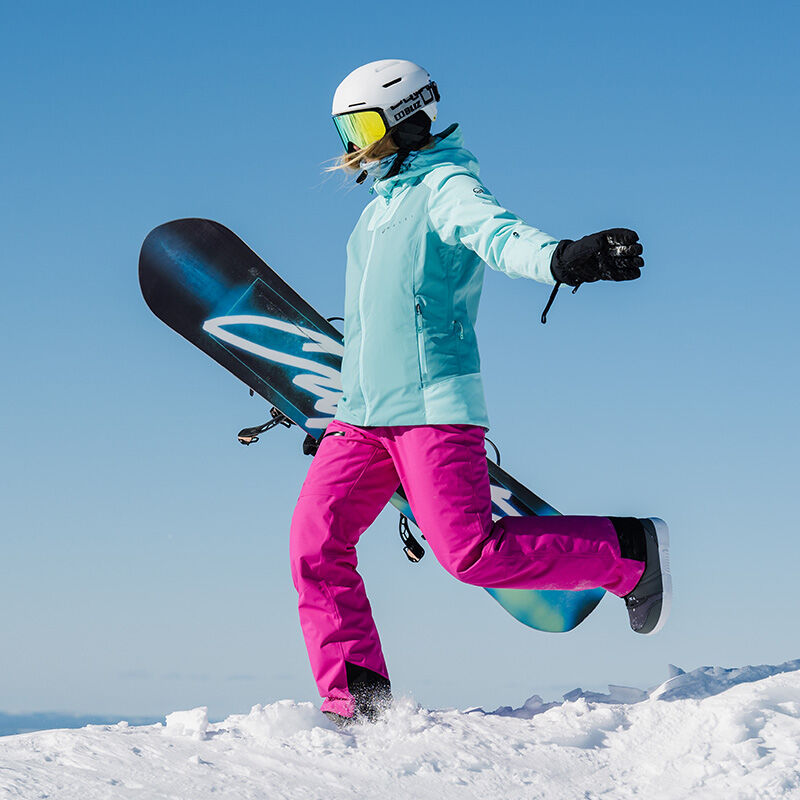 Frau in Halti Carvey DX Skihose hält ein Snowboard und läuft unter blauem Himmel im Schnee.