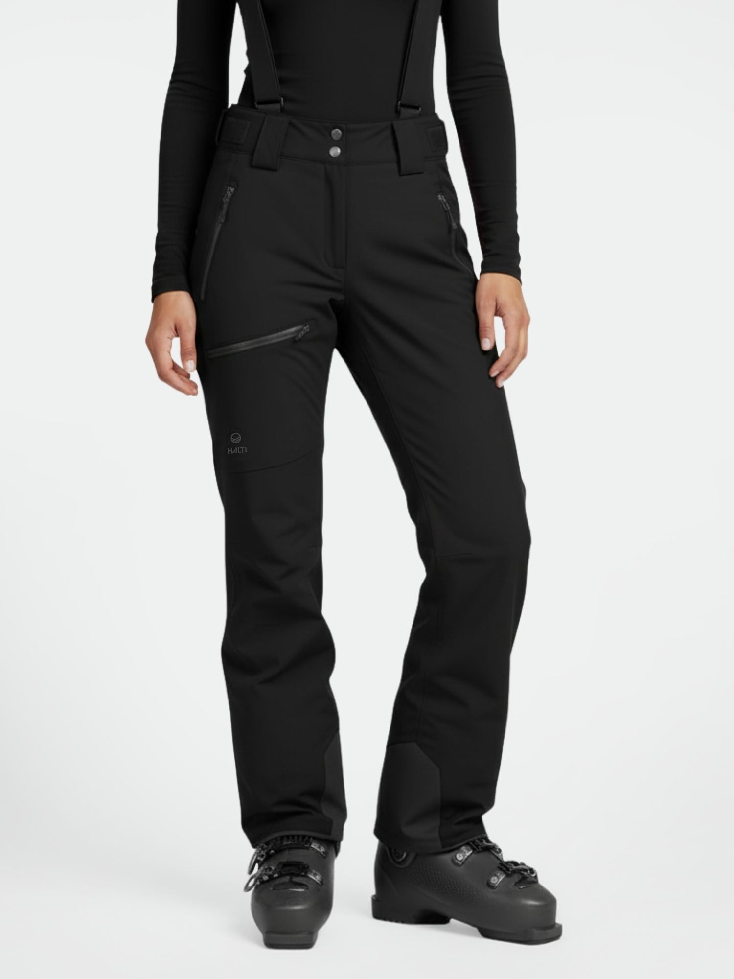 Person in Halti Carvey DX Skihose Damen mit Hosenträgern, schwarzem Top und Skischuhen, aufrecht stehend.