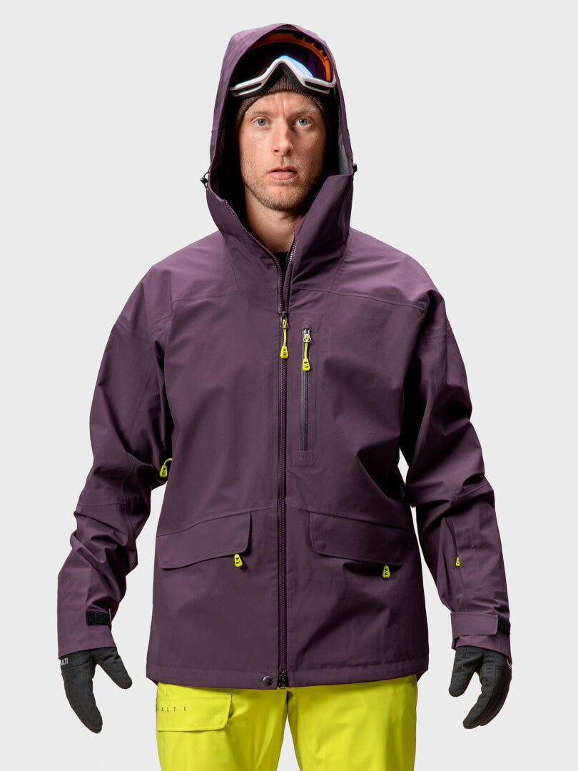 Ein Mann trägt eine Halti Settler 3L DX Ski Jacke Herren in lila, gelbe Hosen, Handschuhe und eine Skibrille vor einem schlichten Hintergrund.