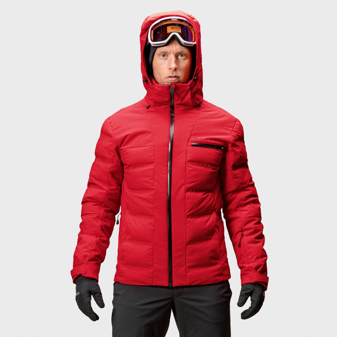 Eine Person in einer roten Winterjacke mit Kapuze, schwarzen Handschuhen und einer Skibrille steht mit dem Gesicht nach vorne vor einem schlichten Hintergrund.
