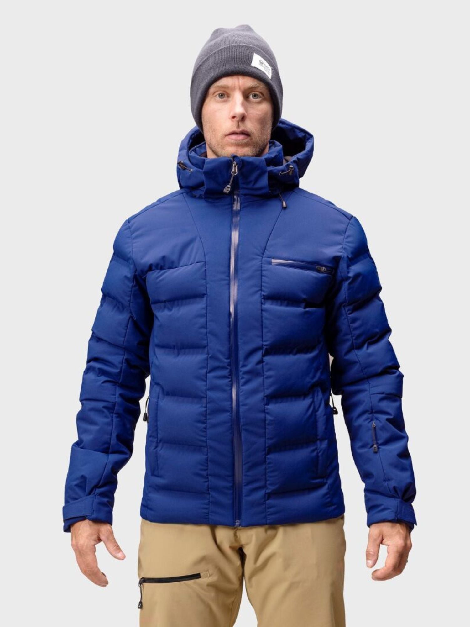 Eine Person in einer blauen Halti Nordic Arcty Skijacke Herren, grauer Mütze und hellbrauner Hose steht mit dem Gesicht nach vorne vor einem schlichten Hintergrund.