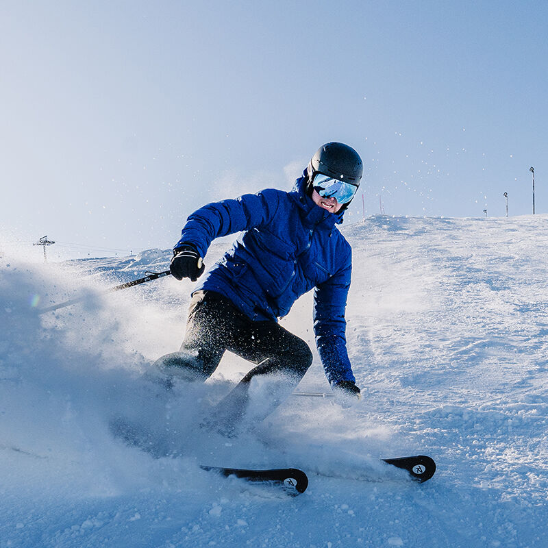 Ein Skifahrer in einer Halti Nordic Arcty Skijacke für Herren und einem Helm fährt bergab, der Schnee fliegt vor einem blauen Himmel.