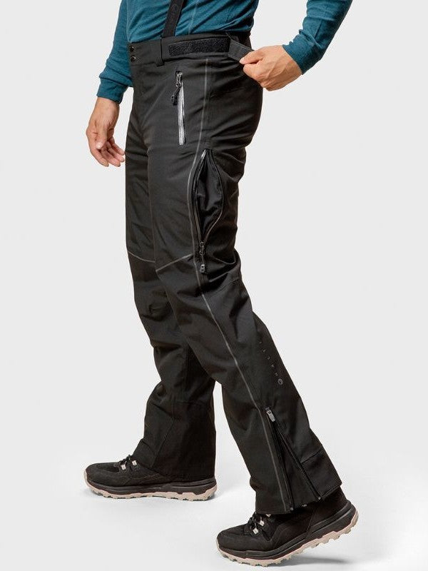 Eine Person trägt Halti Vertica DrymaxX Skihose Herren, ein blaues langärmeliges Hemd und schwarze Wanderschuhe vor einem hellen Hintergrund.