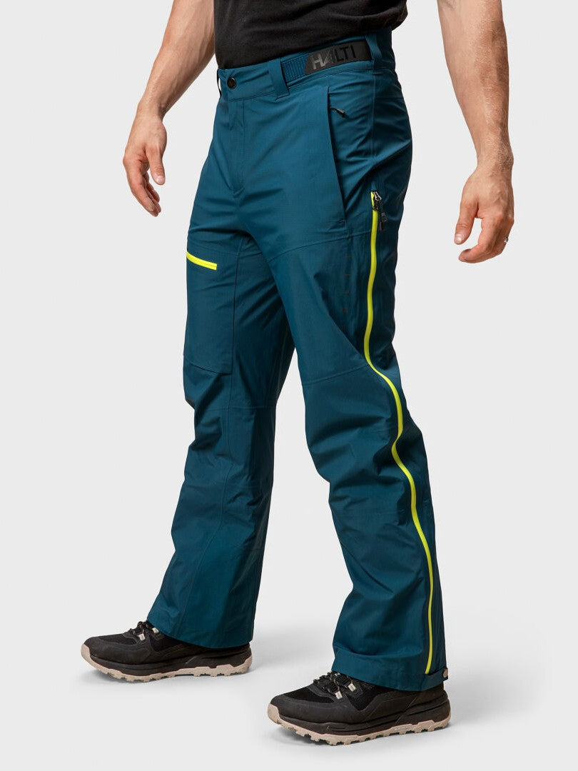 Eine Person in Halti Alpine 3L DX Skihose Herren teal Hosen mit gelben Reißverschlüssen und schwarze Wanderschuhe, auf einem unifarbenen Hintergrund.