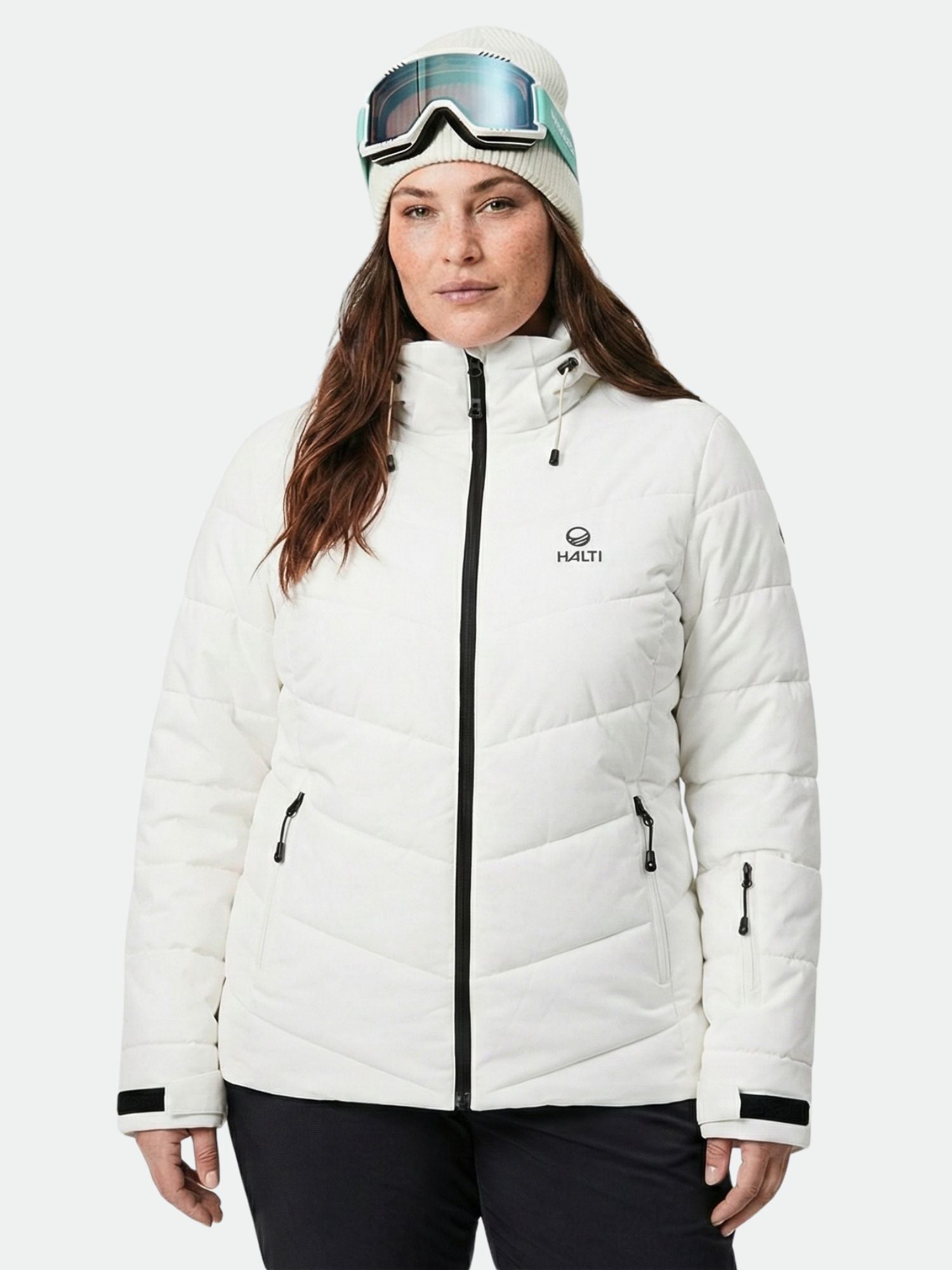 Eine Frau in einer Halti Mellow Puffer Skijacke Damen Plus, einer schwarzen Hose, einer weißen Mütze und einer Skibrille schaut nach vorne.