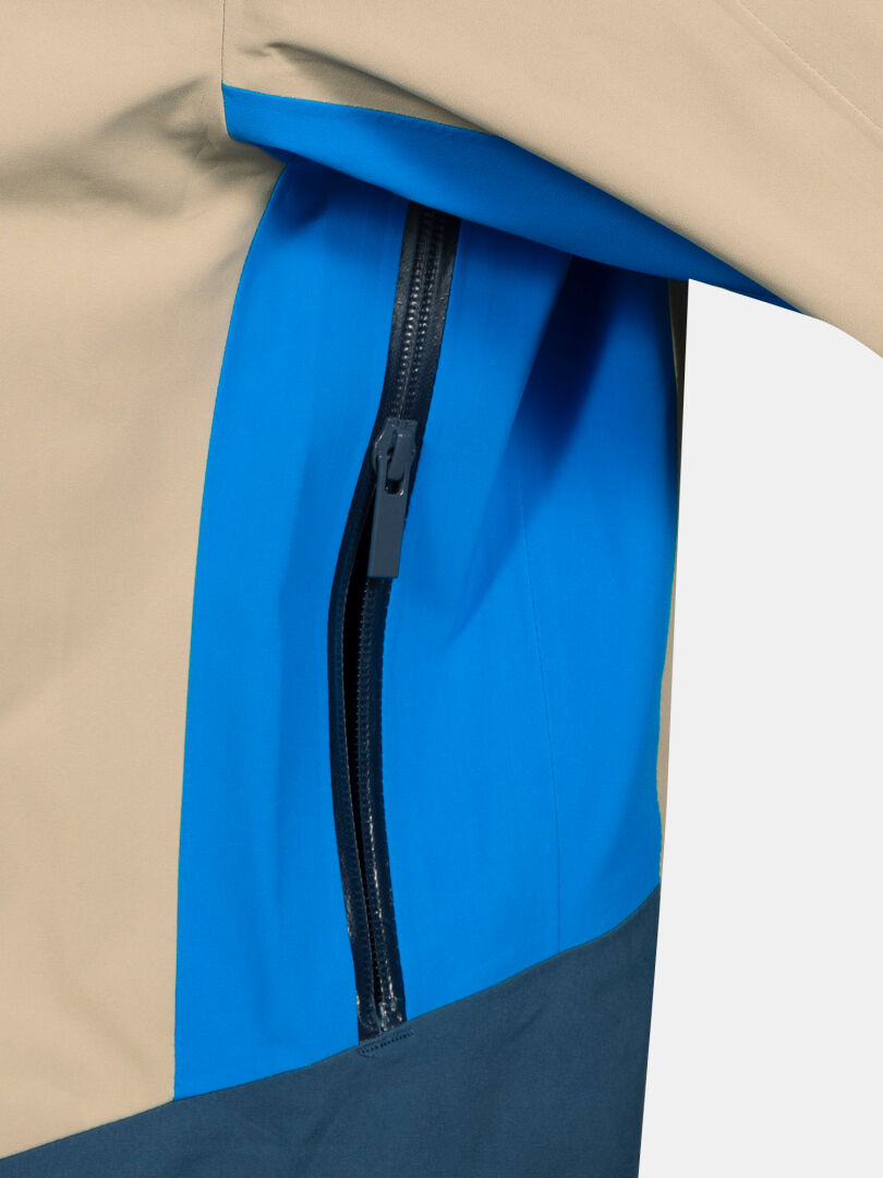 Nahaufnahme der Halti Rizle Unisex 3L DX Skijacke mit einer vertikalen Reißverschlusstasche und blau-beigem Farbblocking.