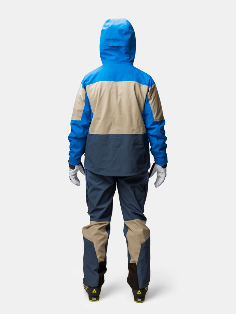 Person in einer Halti Rizle Unisex 3L DX Skijacke und Hose, blau/beige/dunkelgrau, stehend mit dem Gesicht zur Seite auf einfarbigem Hintergrund.