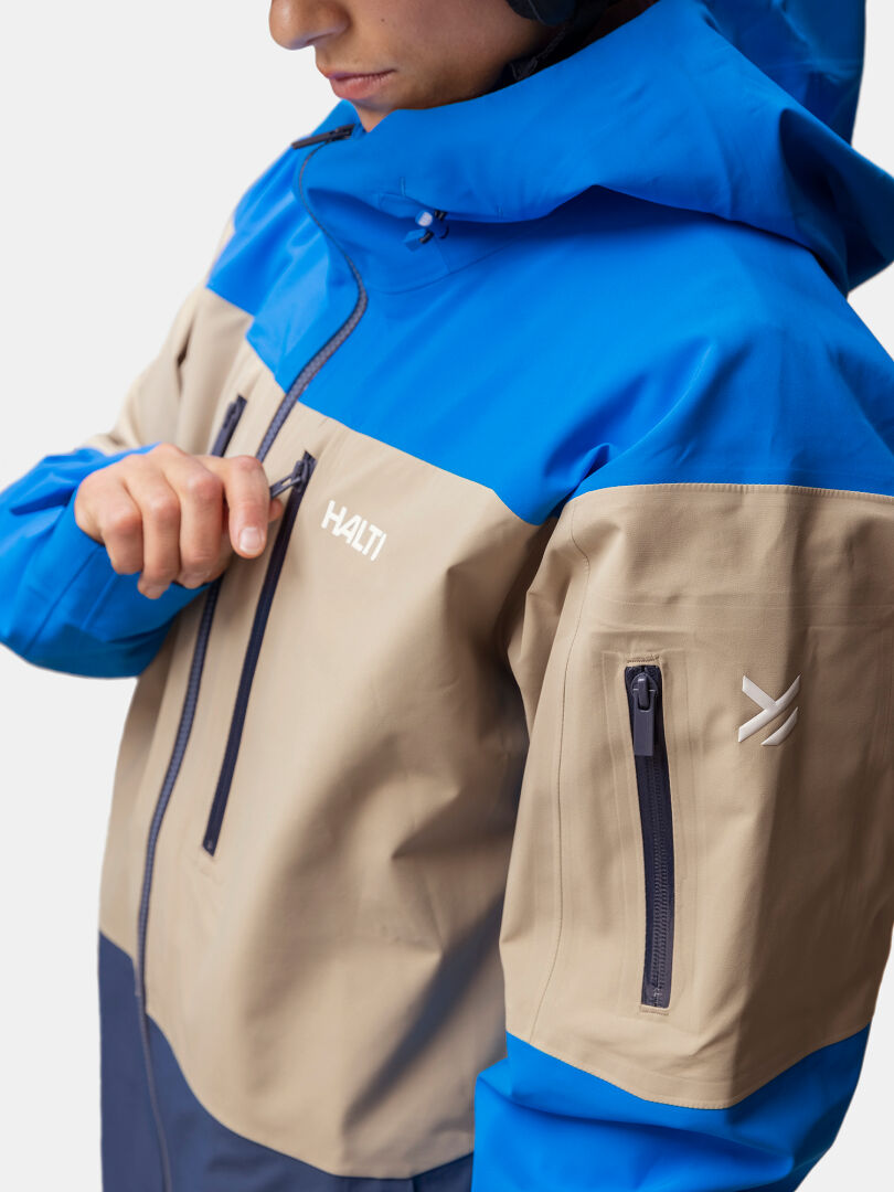 Eine Person in einer blau-beigen Halti Rizle Unisex 3L DX Skijacke öffnet eine Brusttasche; eine Armtasche mit Reißverschluss ist sichtbar.