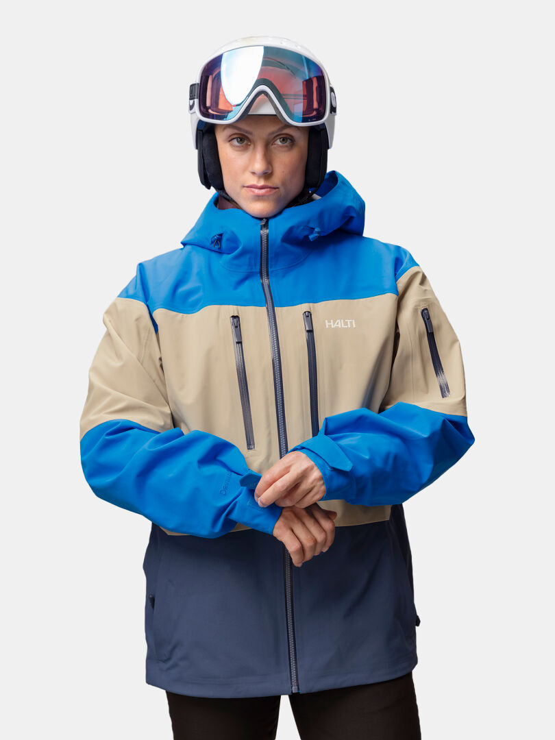 Eine Person trägt eine blaue, beige und graue Halti Rizle Unisex 3L DX Skijacke mit DrymaxX Nano tech, eine Skibrille und einen Helm.