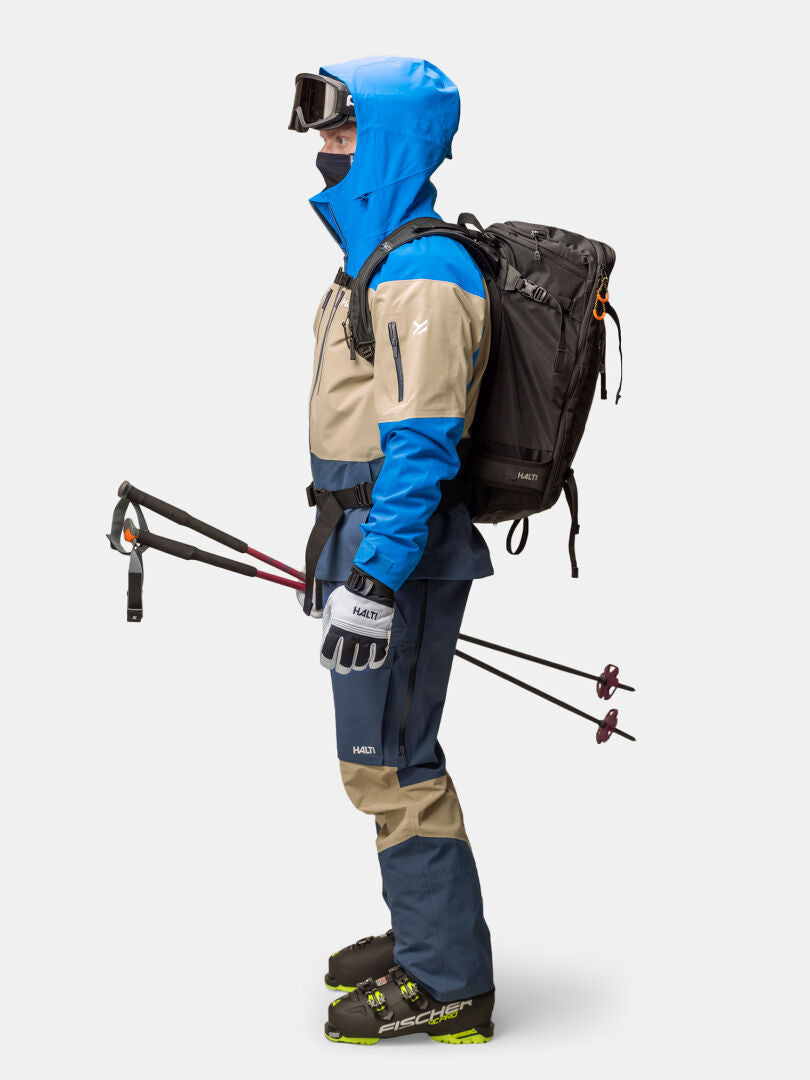 Eine Person in einer Halti Rizle Unisex 3L DX Skijacke mit Skibrille, Handschuhen, Rucksack und Skistöcken, stehend im Profil.