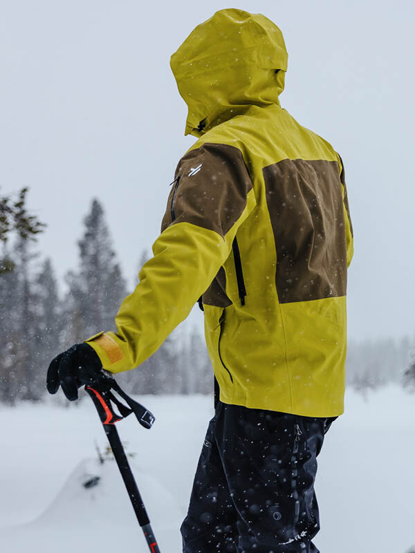 Eine Person in einer Halti Rizle Unisex 3L DX Skijacke steht mit einer Stange in der Hand im Schnee, mit Bäumen und fallendem Schnee dahinter.