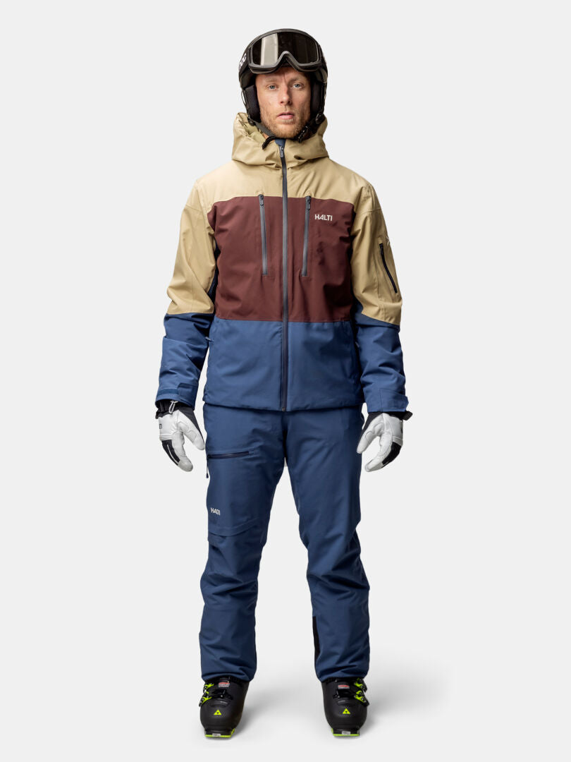 Eine Person steht vorne und trägt die Halti Melvik DX Skijacke Herren in Beige, Braun und Blau sowie eine komplette Skiausrüstung.