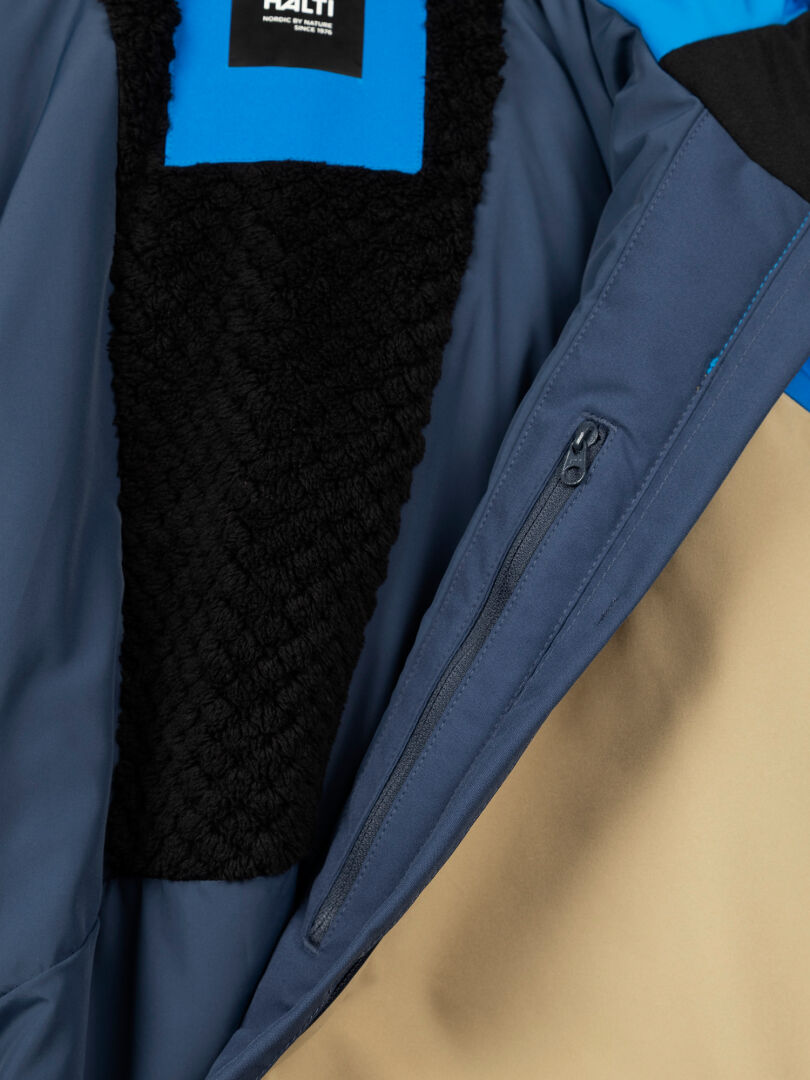 Nahaufnahme einer Halti Melvik DX Skijacke Herren mit blauem, beigem und schwarzem Stoff, einer Reißverschlusstasche und einem Etikett am Kragen.