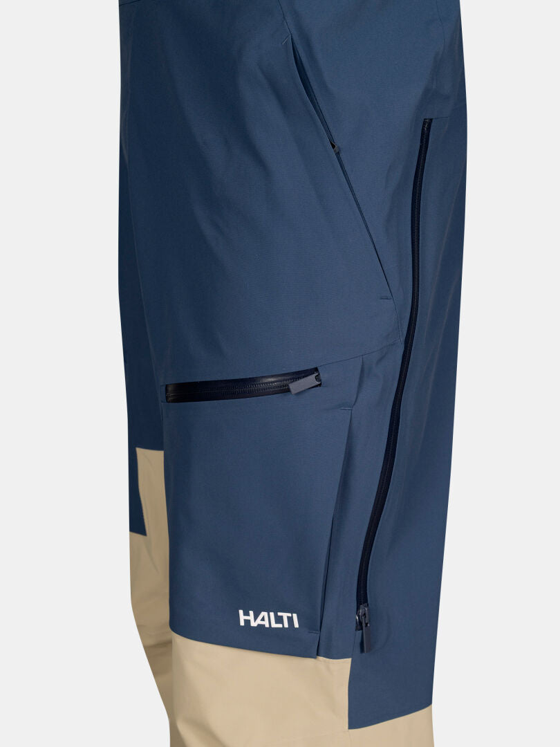 Nahaufnahme der Rizle 3L DX Skihose Unisex von Halti in Blau und Beige mit Reißverschlusstaschen und Logo in Kniehöhe.