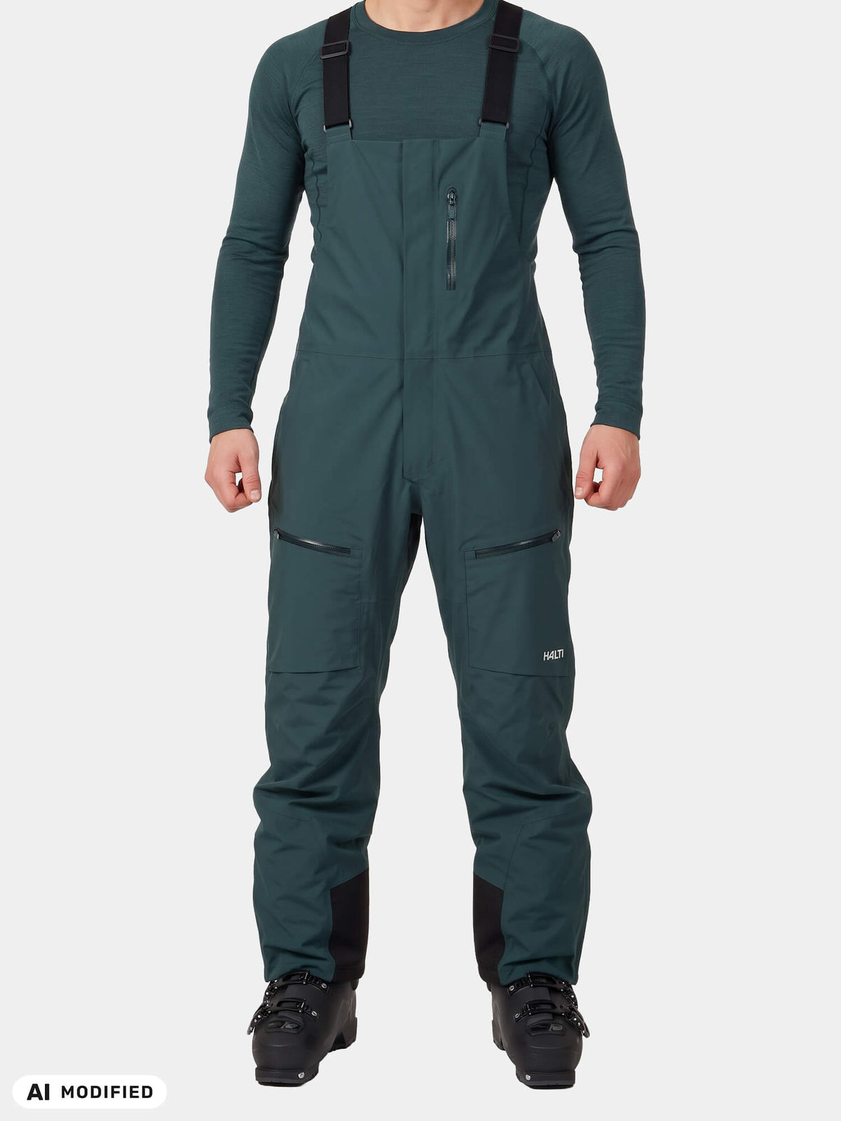 Eine Person trägt dunkelgrüne, wasserdichte Halti Rizle 3L DX Skihosen – eine Unisex-Latzhose mit schwarzen Hosenträgern – sowie Skischuhe.