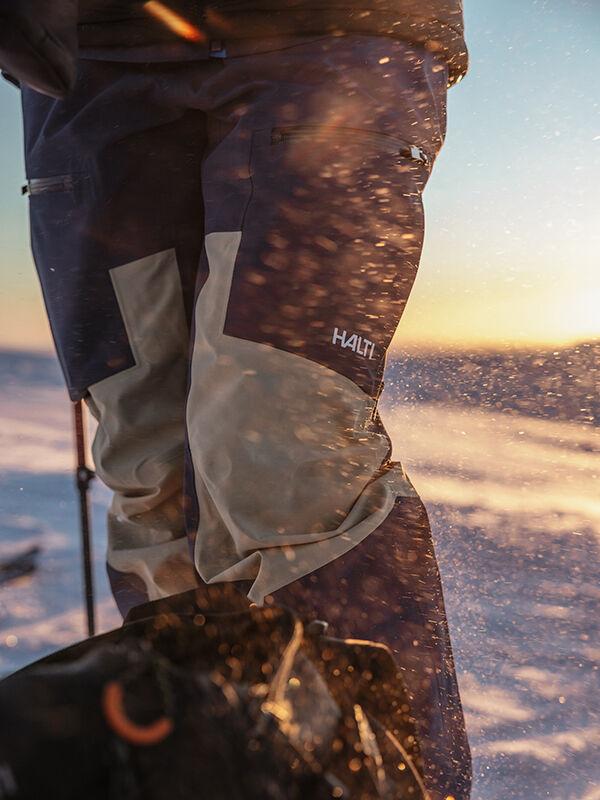 Eine Person in Halti Rizle 3L DX Skihose Unisex steht in einer verschneiten Landschaft mit Sonnenlicht und aufgewirbelten Schneepartikeln.