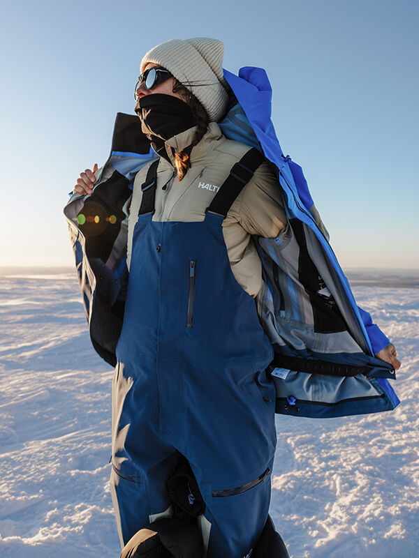 Eine Person in Winterkleidung zieht ihre Halti Rizle 3L DX Skihose Unisex in einer verschneiten Landschaft unter klarem Himmel an.