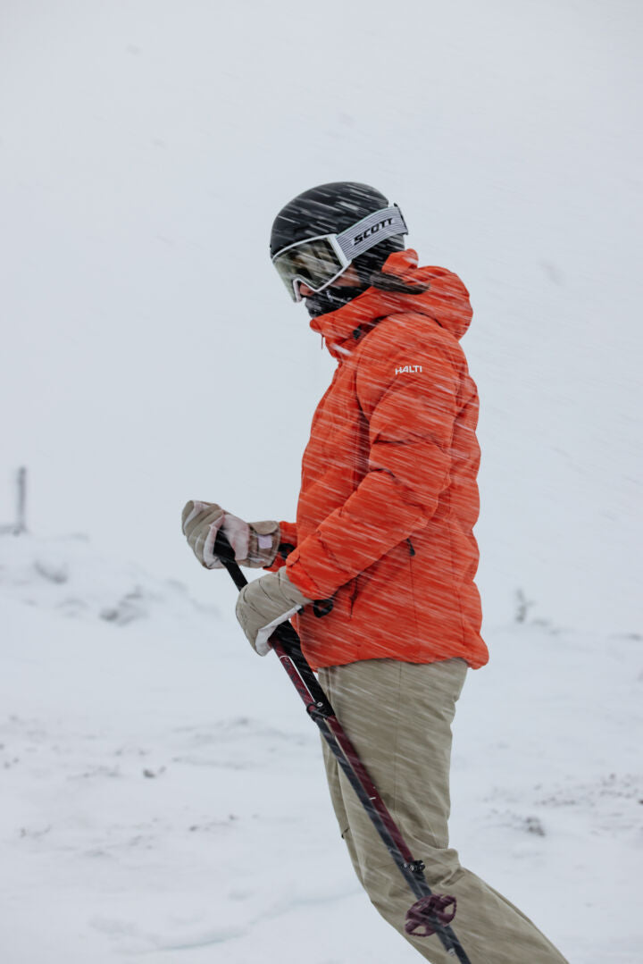 Person in orangefarbener Jacke und Helm hält Skistöcke im Schneewind, trägt Halti Carvey II DX Skihose Damen.