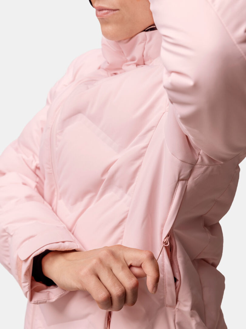 Eine Frau in einer hellrosa Halti Nordic Arcty II Skijacke öffnet den Reißverschluss einer Achselhöhle, wobei ihr Gesicht teilweise sichtbar ist.