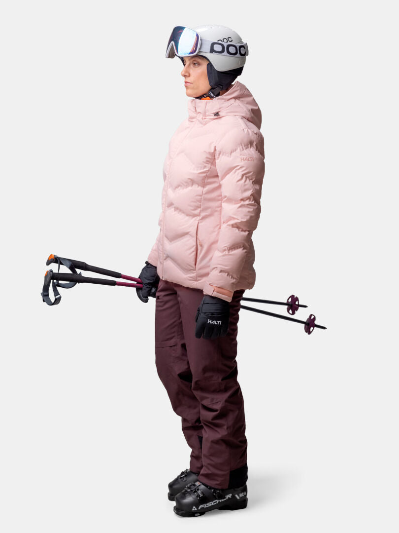 Eine Person in Skiausrüstung hält die Skistöcke und trägt die Halti Nordic Arcty II Skijacke Damen, eine kastanienbraune Hose, einen Helm, Handschuhe und eine Skibrille.