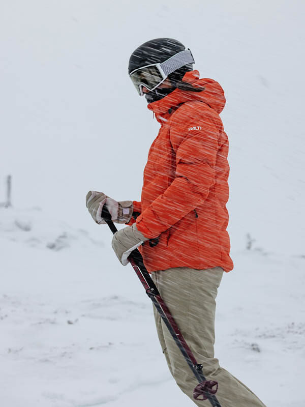 Eine Frau in einer Halti Nordic Arcty II Skijacke steht mit Skibrille und Stöcken in einer verschneiten, windigen Landschaft.