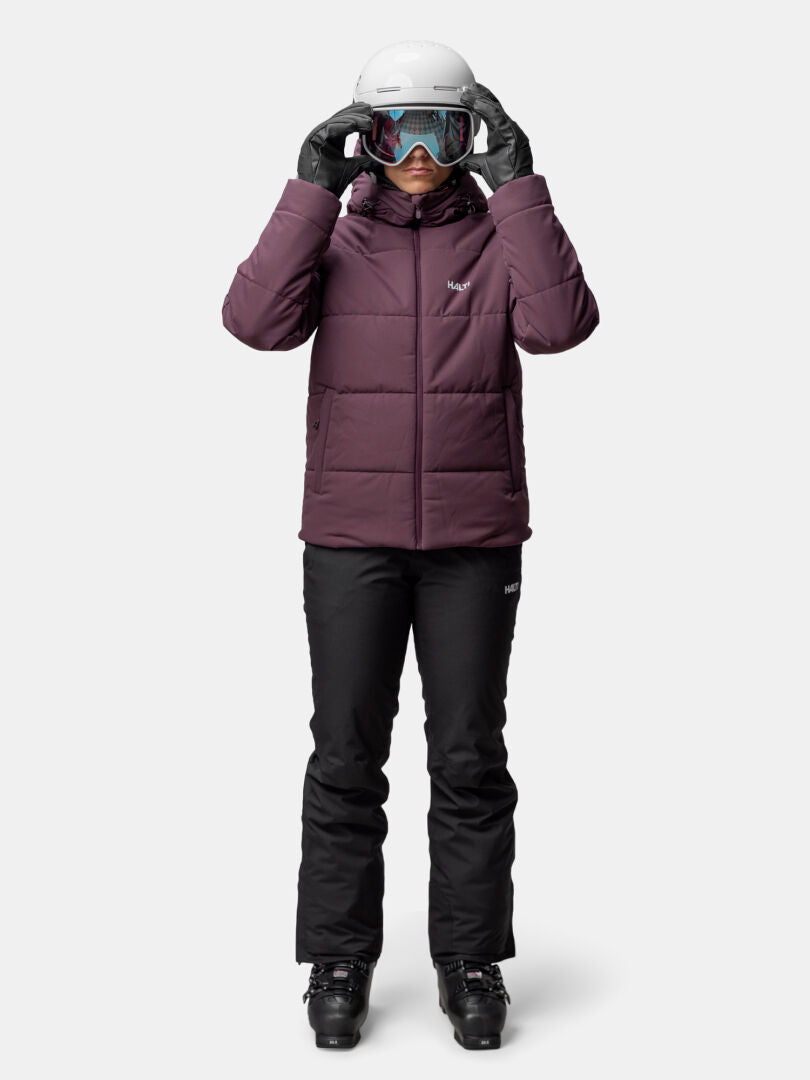 Frau in Halti Ascale Puffer Skijacke Damen, schwarze Skihose, Skihelm und Skibrille vor neutralem Hintergrund.
