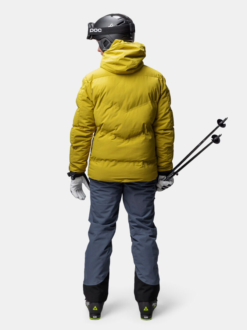 Person in einer Halti Nordic Arcty II Skijacke Herren, graue Hose, Helm, und Handschuhe steht mit Skistöcken, Rückenansicht.
