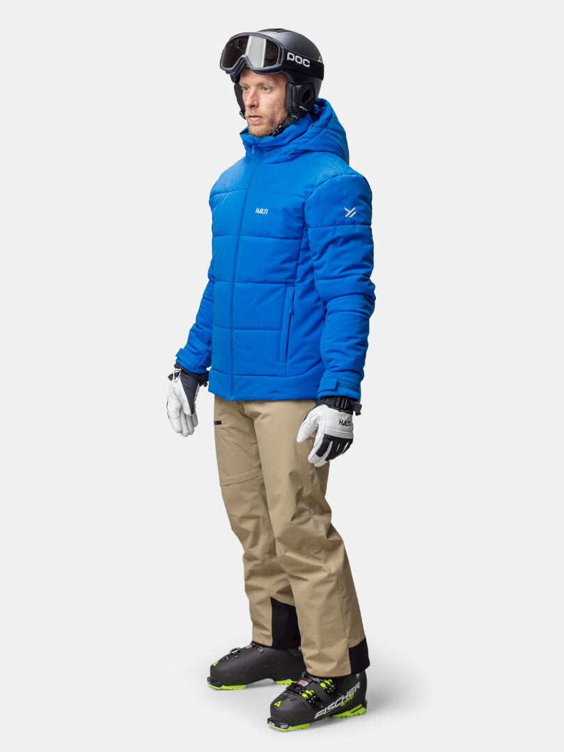 Eine Person in einer Halti Ascale Puffer Skijacke Herren, beiger Hose, schwarzen Skistiefeln, weißen Handschuhen und einem schwarzen Helm mit Brille.